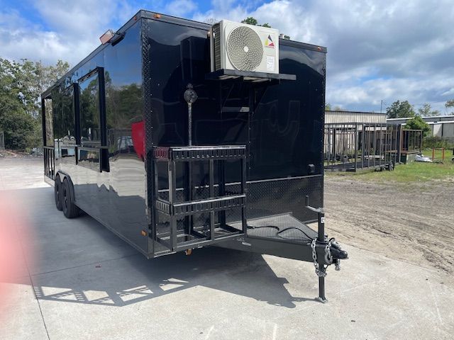 Black food trailer with serving windows, air conditioning unit, and metal stairs.