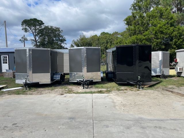 Cargo trailers parked outdoors on a sunny day. Silver, black, and white trailers are side by side.