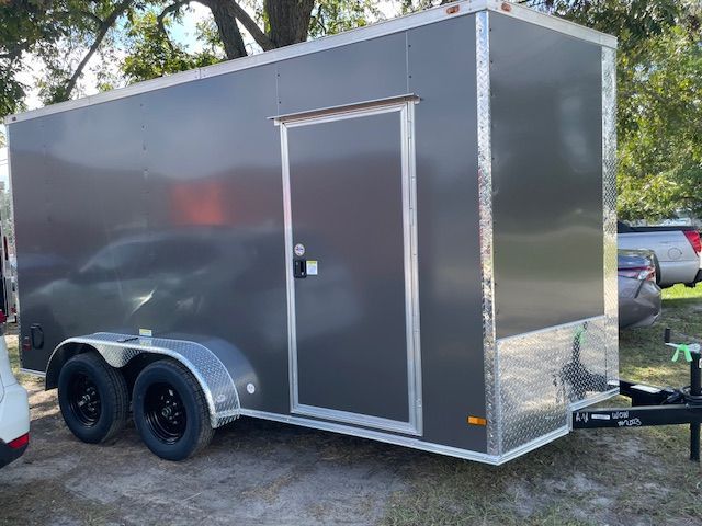 A gray enclosed cargo trailer with a side door, parked outdoors with vehicles visible.