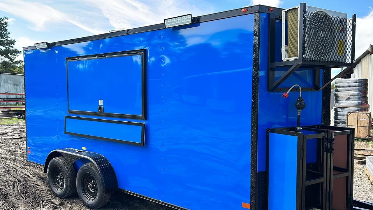Blue food trailer with serving window, black wheels, and an exterior refrigerator.