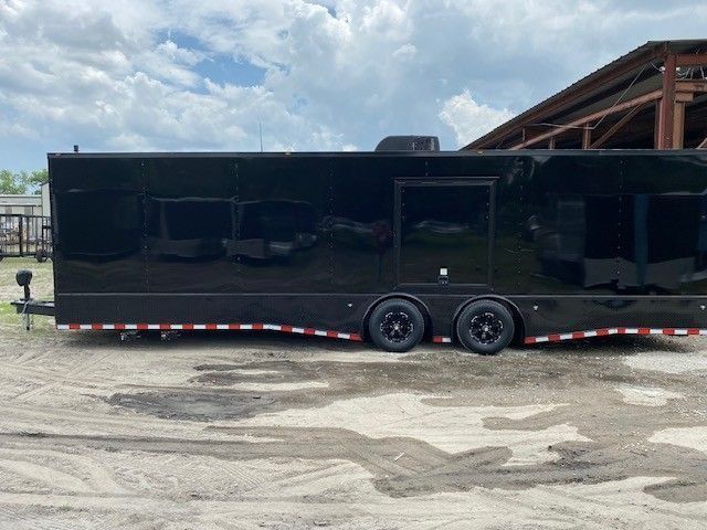 Black enclosed trailer with red and white trim, sitting on a dirt surface under a cloudy sky.