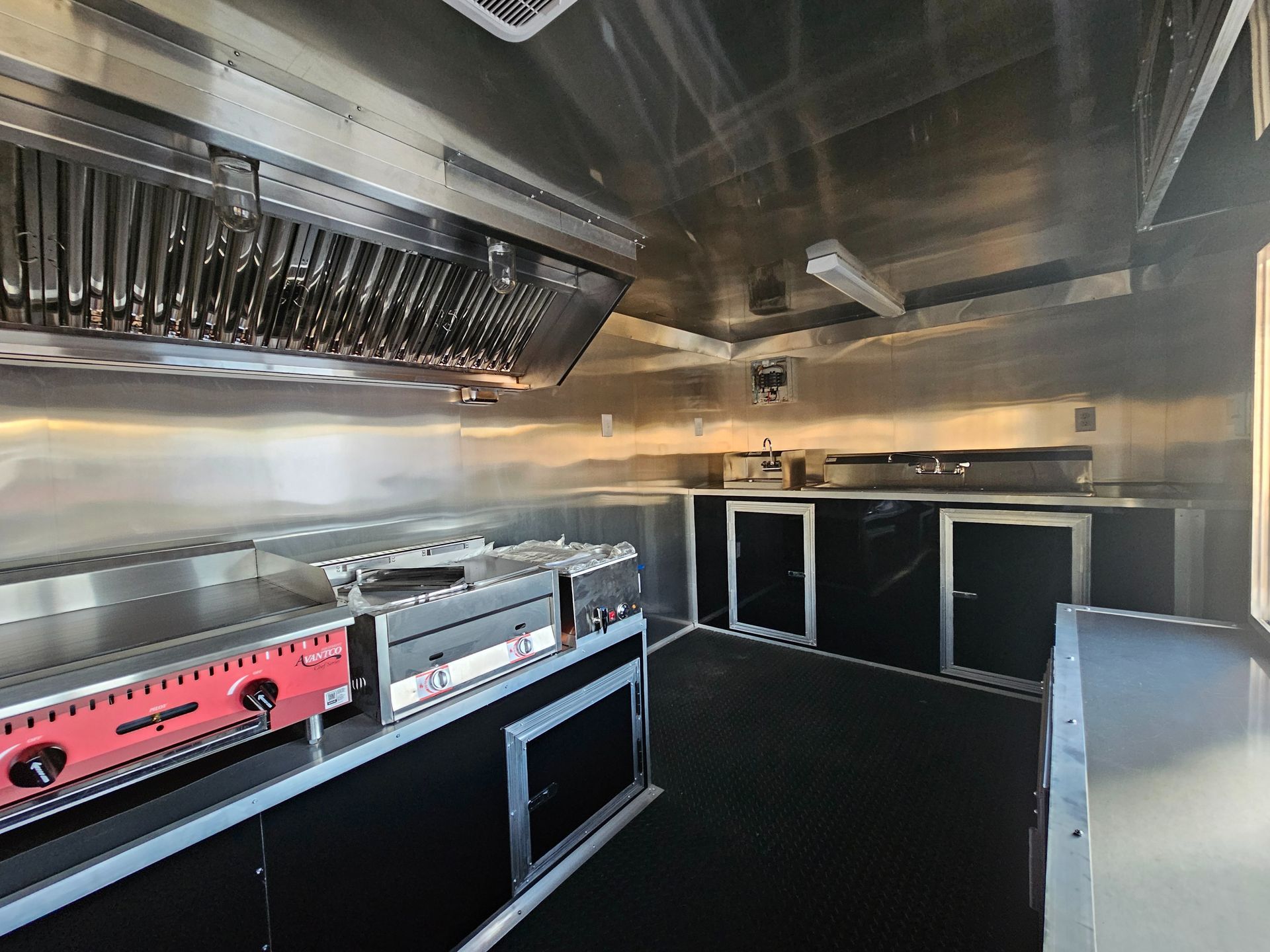 Interior of a food truck kitchen: stainless steel counters, appliances, range, exhaust hood, and black cabinets.