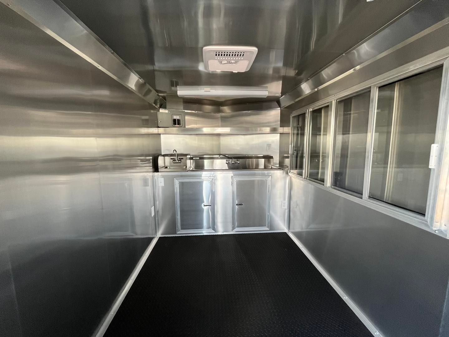 Interior of a food truck, stainless steel walls, black flooring, windows on the right, and a kitchen area in the back.
