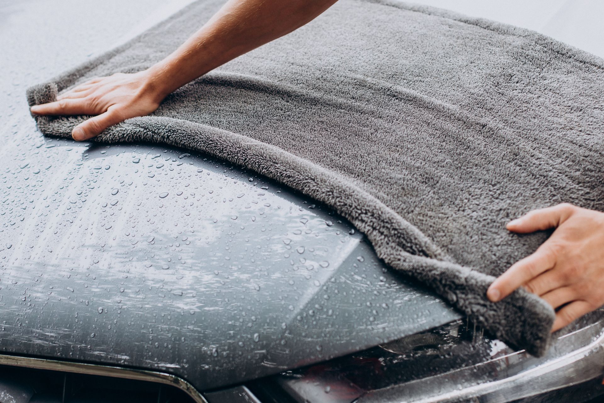 Hands wiping soap and water off a car windshield with a gray microfiber towel