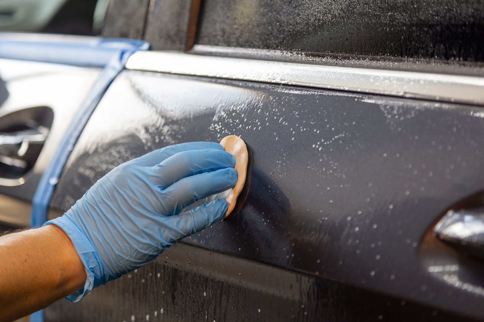 Gloved hand wiping a black car door with a sponge during car cleaning