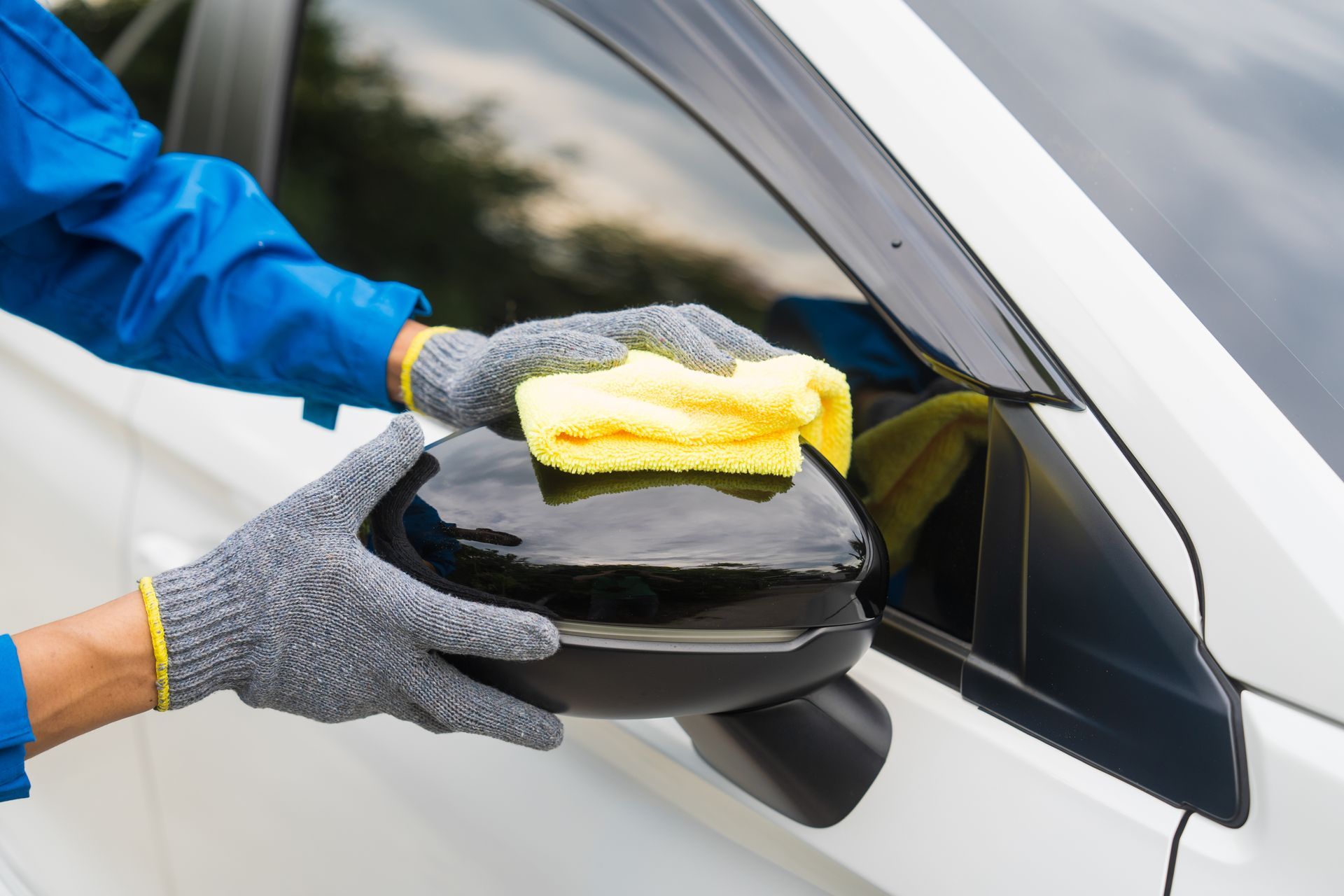 Gloved hands cleaning a white car’s side mirror with a yellow cloth and black sponge