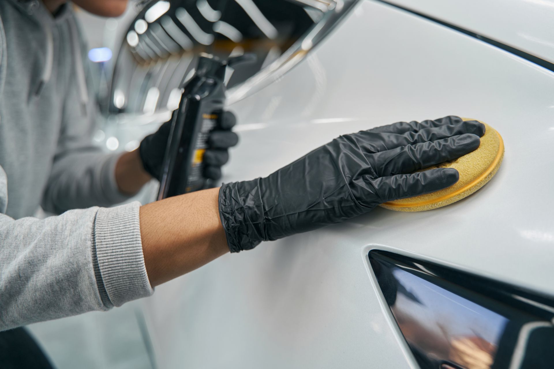 Gloved hand polishing a white car with a yellow buffing pad in an auto shop