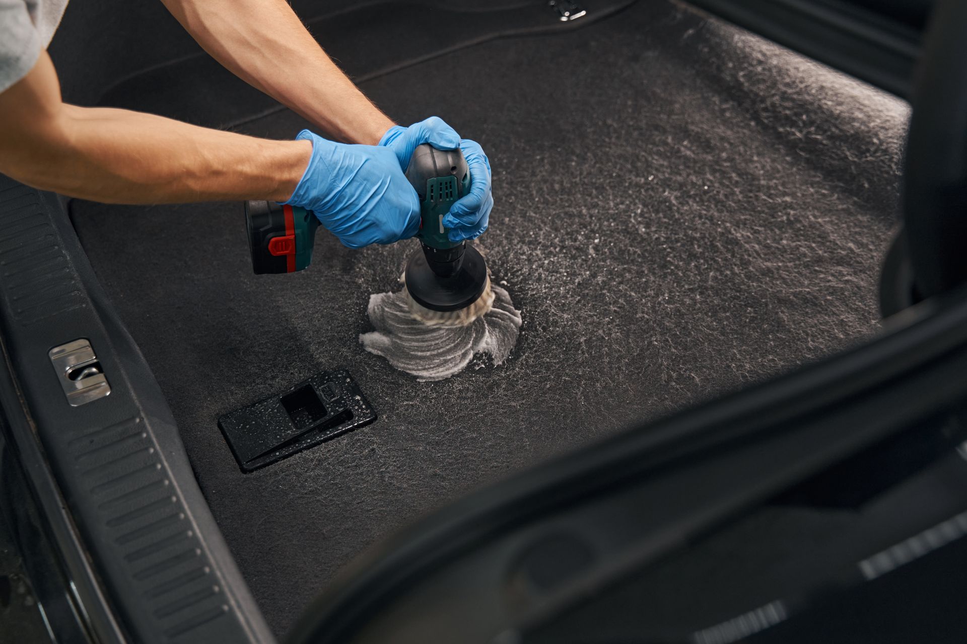 Gloved hands cleaning a car trunk carpet with a handheld vacuum attachment