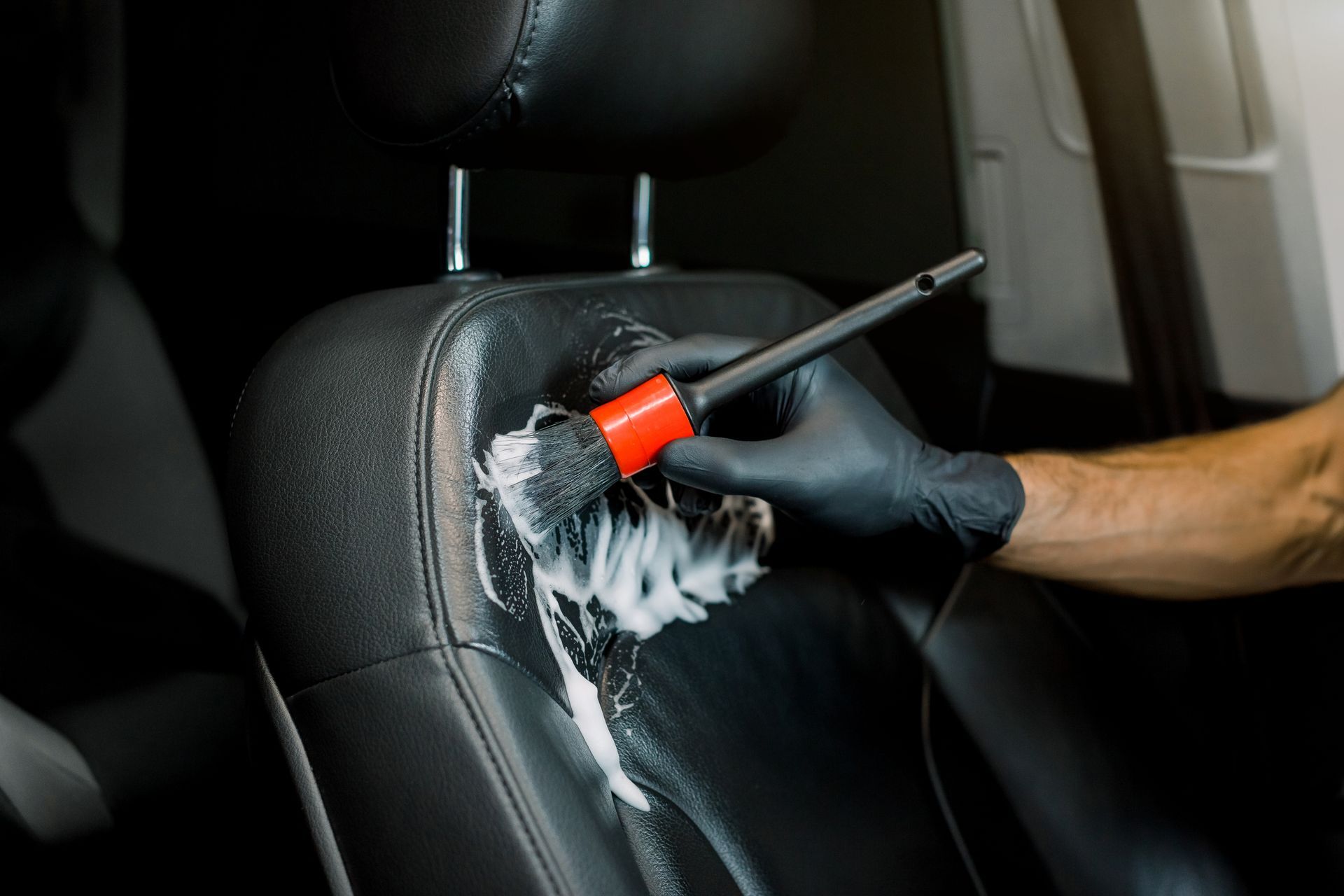 Gloved hand cleaning a black car seat with a brush and foam inside the vehicle