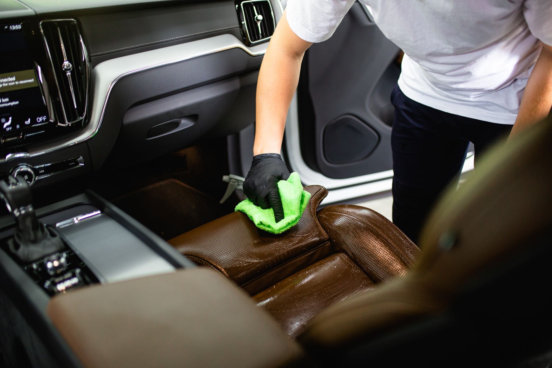 Person cleaning a brown leather car seat with a green cloth inside a vehicle.