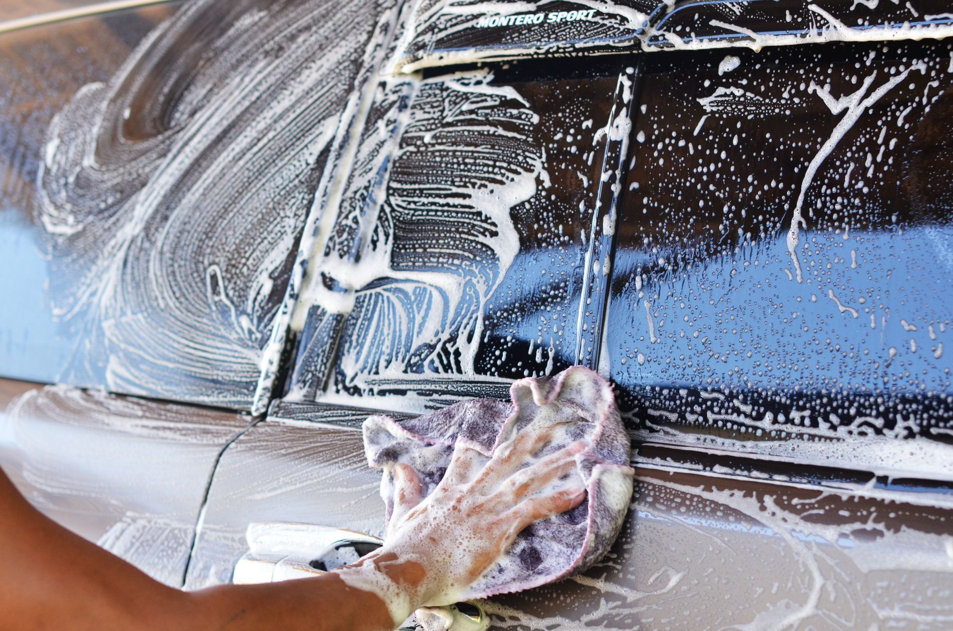 Person washing a dark car door with a soapy sponge, covered in foam and water.