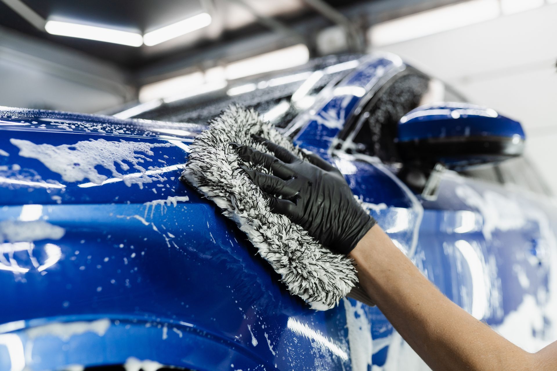 Hand washing a blue car with a gray microfiber mitt in a bright auto shop