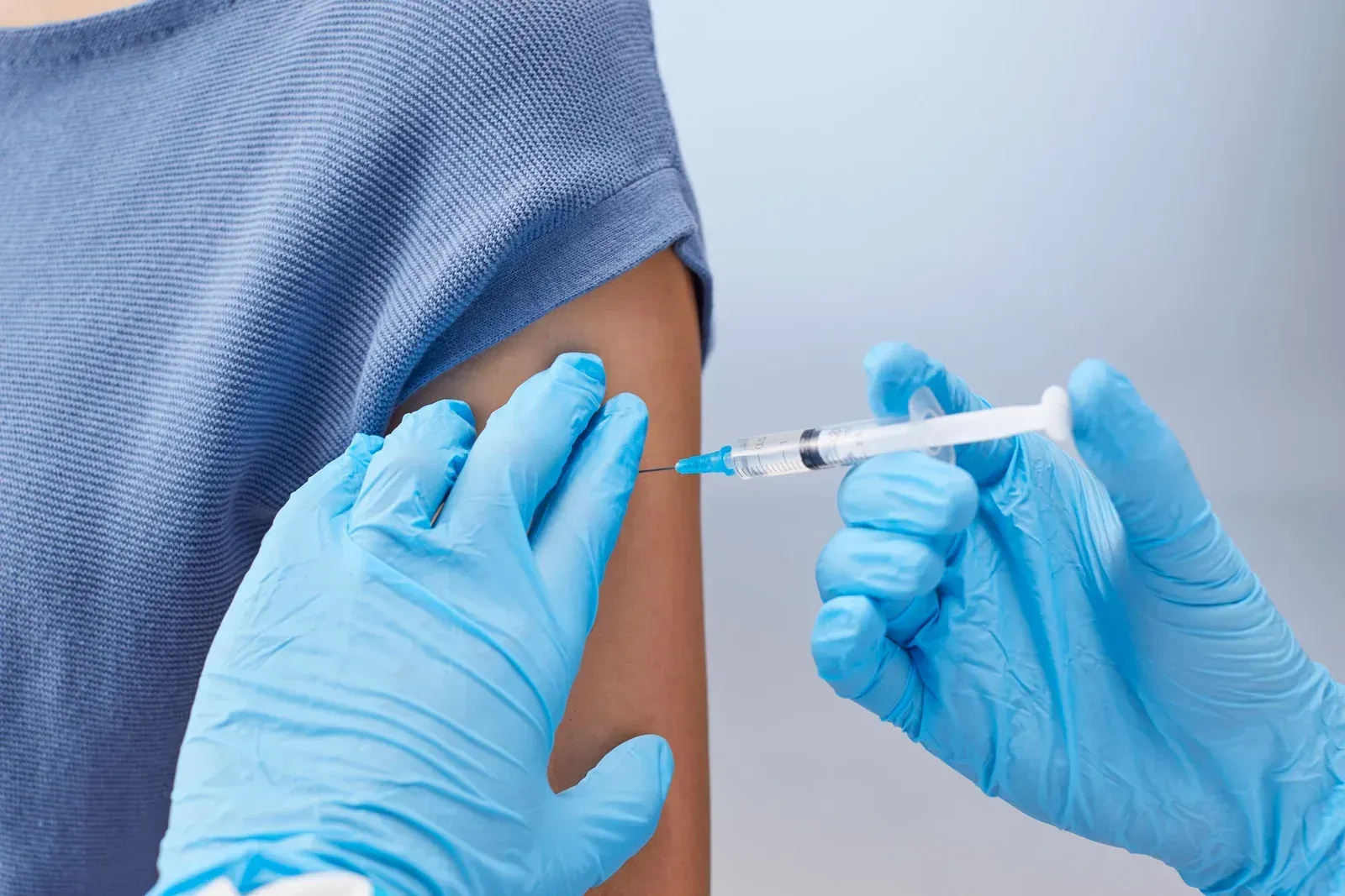 Person receiving a vaccine injection in the arm, blue gloved hands, light blue shirt.