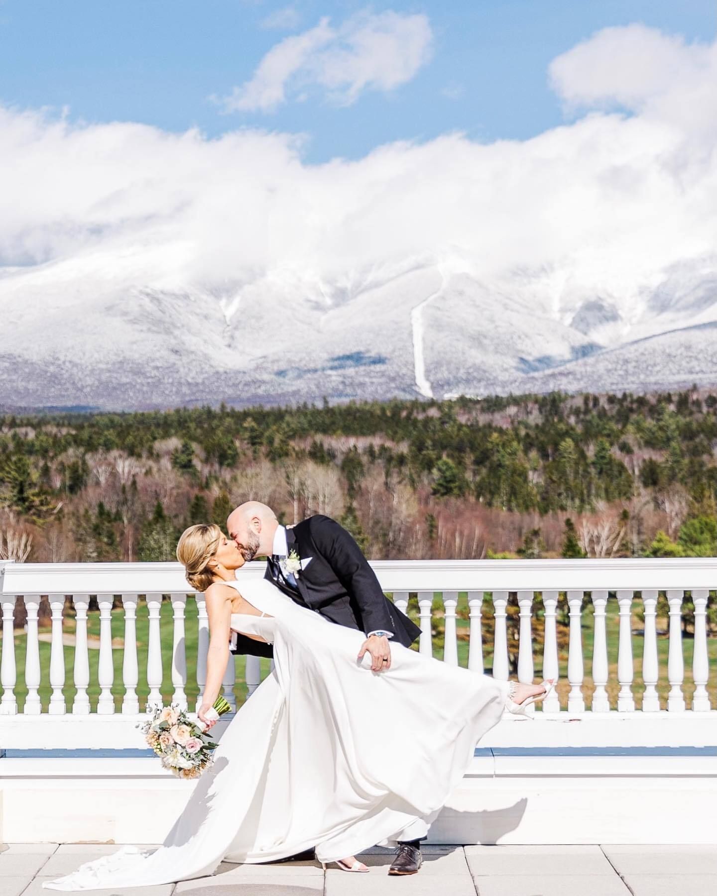 A bride and groom are kissing on a balcony with mountains in the background.