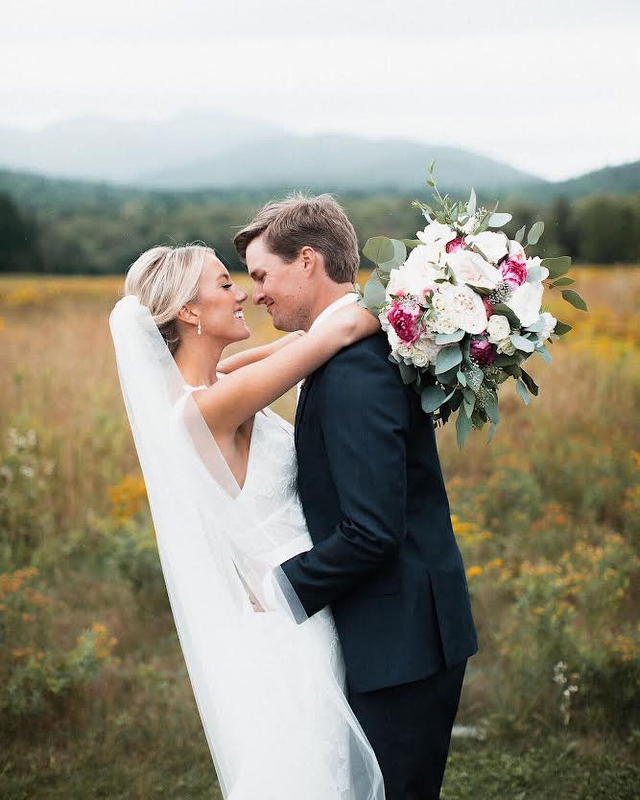 A bride and groom are kissing in a field while the bride is holding a bouquet of flowers.