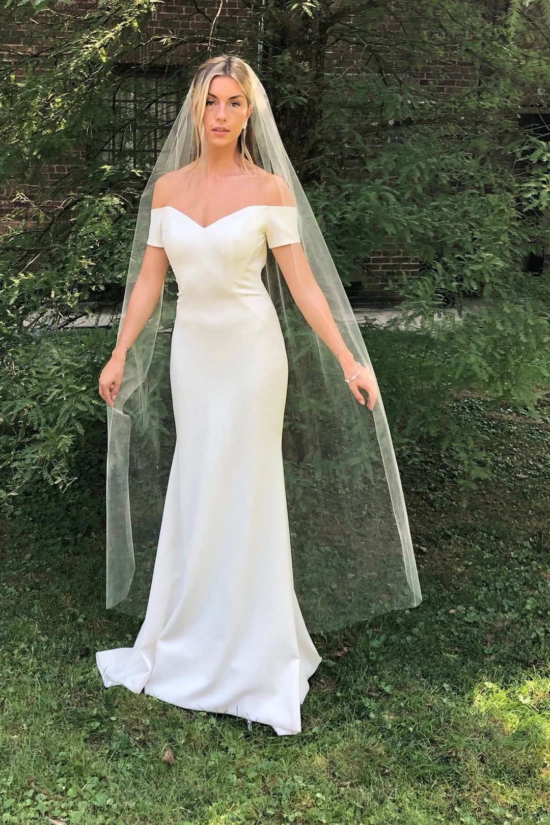 A woman in a white wedding dress and veil is standing in the grass.