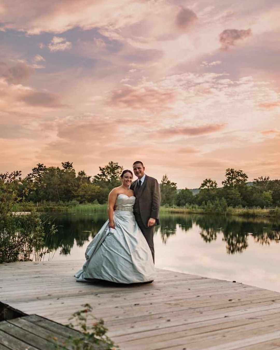 A bride and groom are standing on a dock next to a lake at sunset.