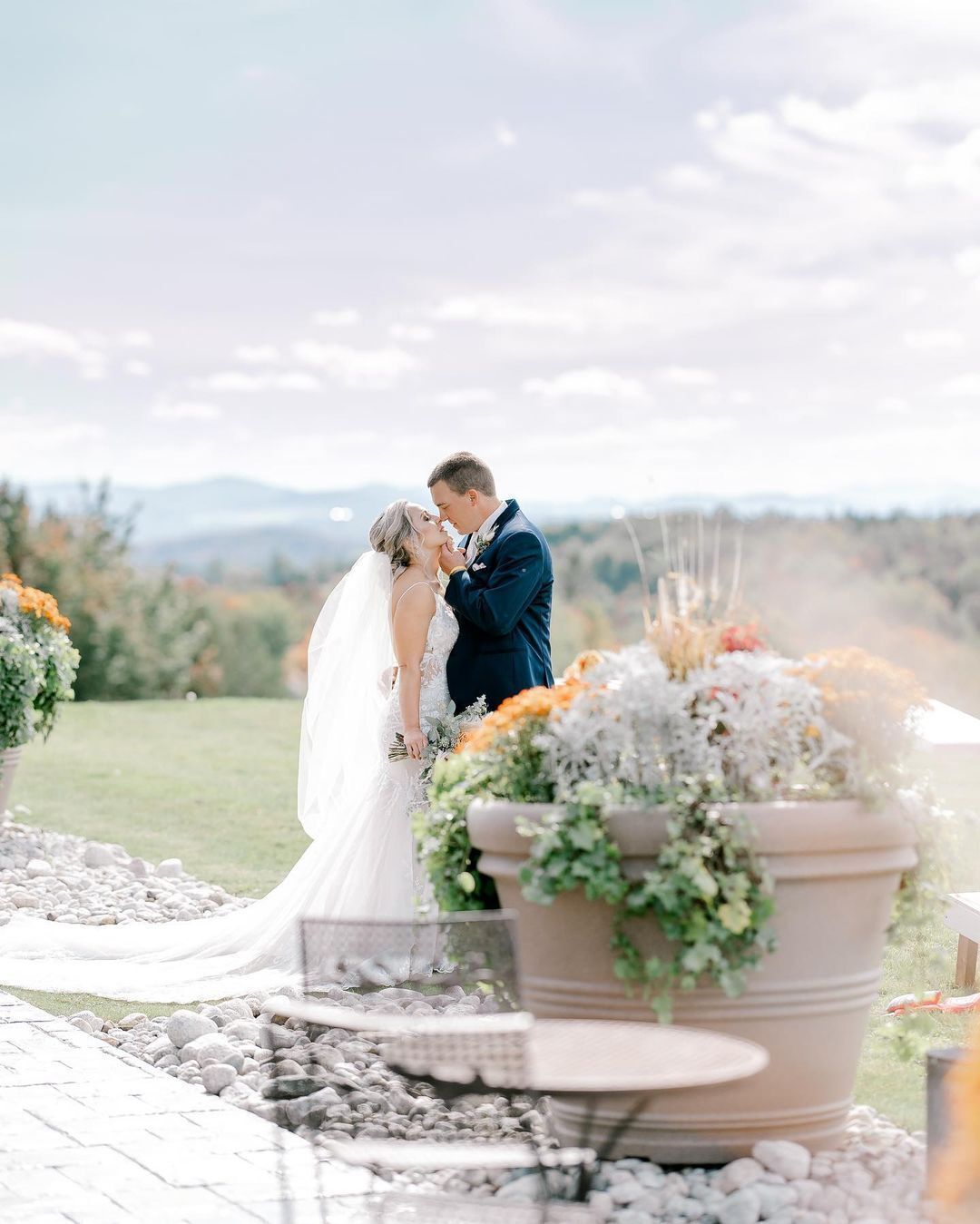 A bride and groom are kissing in front of potted plants.