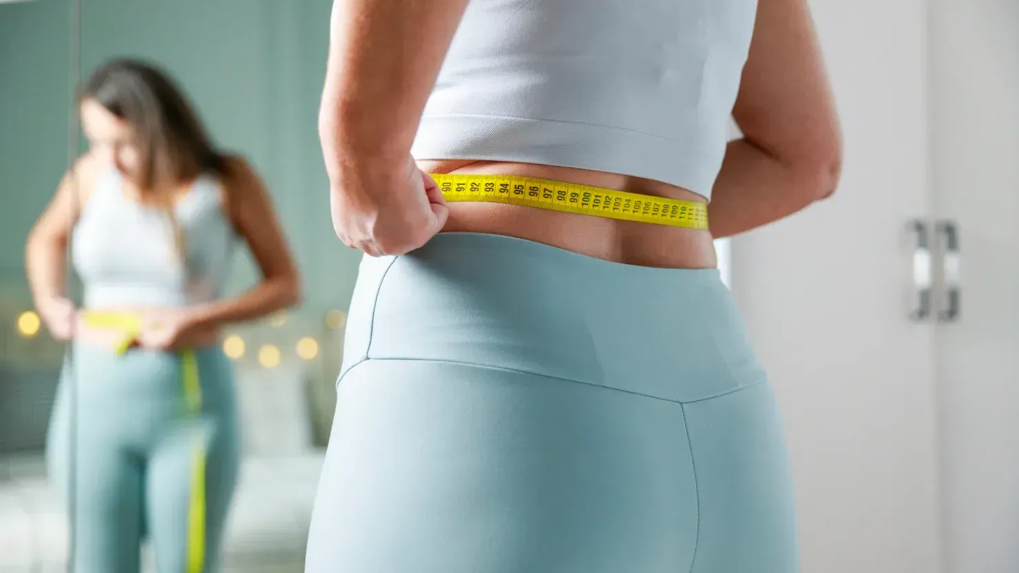 Vrouw meet haar taille met een geel meetlint; weerspiegeling in de spiegel.