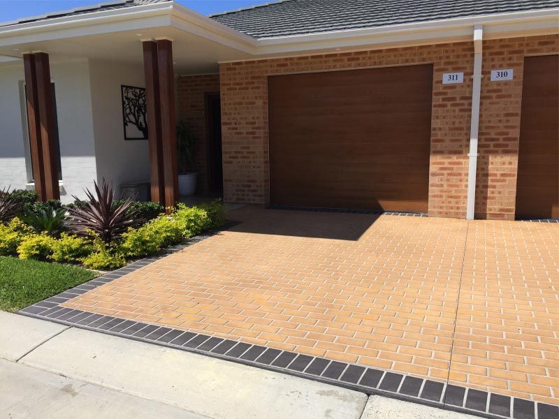 Tan brick driveway leads to a garage with a wooden door. Landscaping with green plants and dark border. — Easy Mix Concrete In Berkeley Vale, NSW