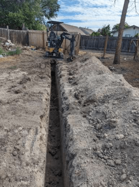 A small excavator is digging a trench in the dirt in a backyard.
