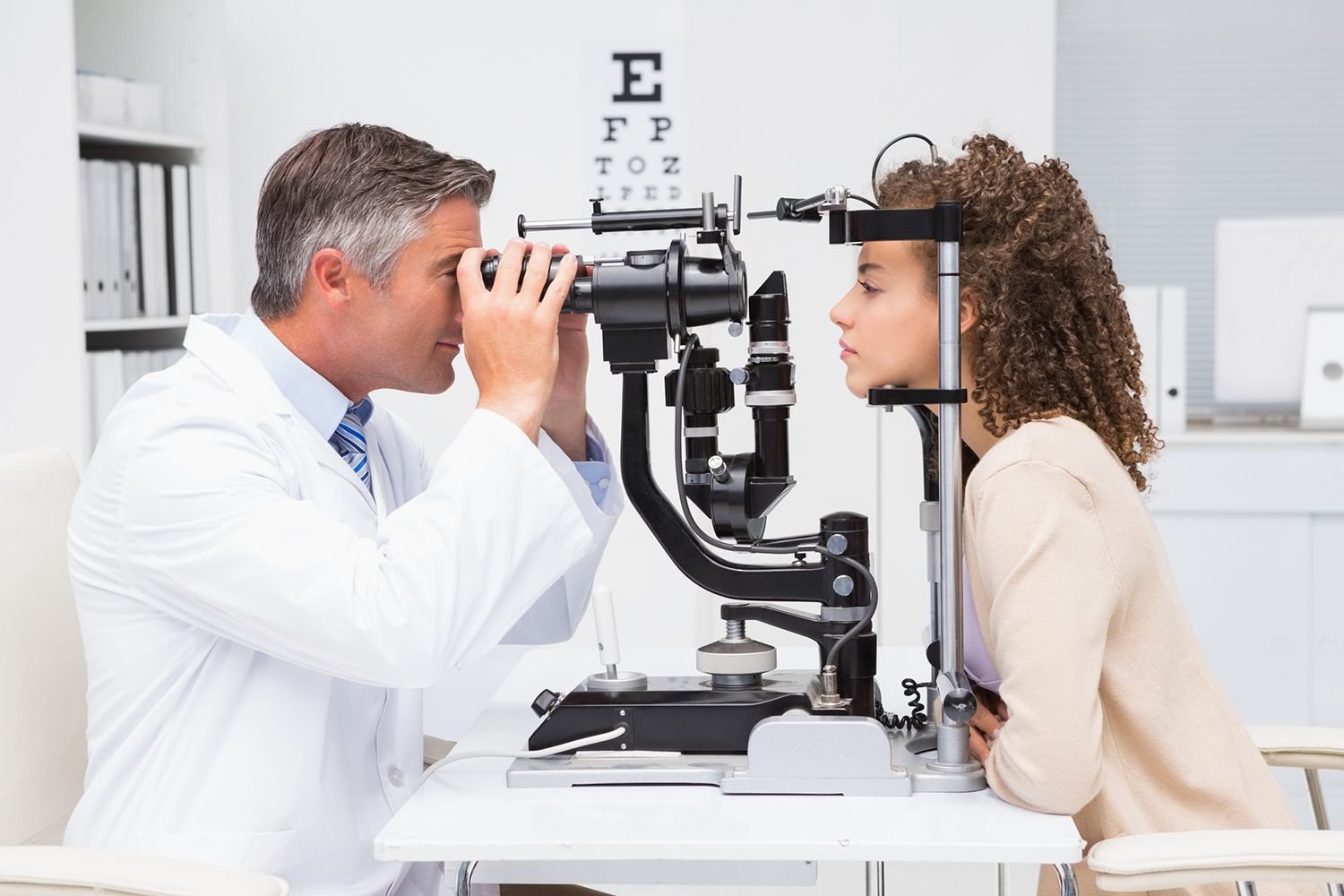 Optometrist performing eye exam with slit lamp microscope in a bright clinical setting.