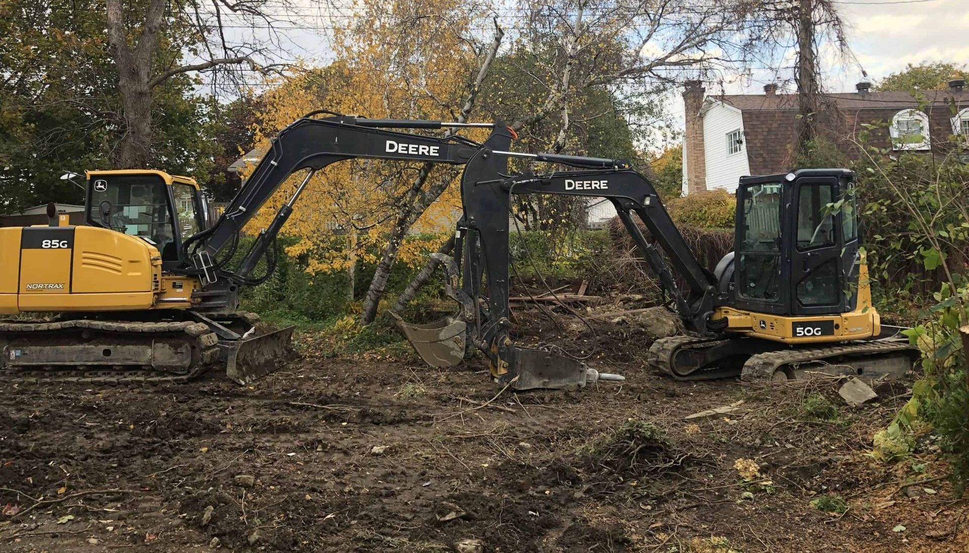 Deux excavatrices dégagent les broussailles et les débris dans une cour boueuse près d'une maison