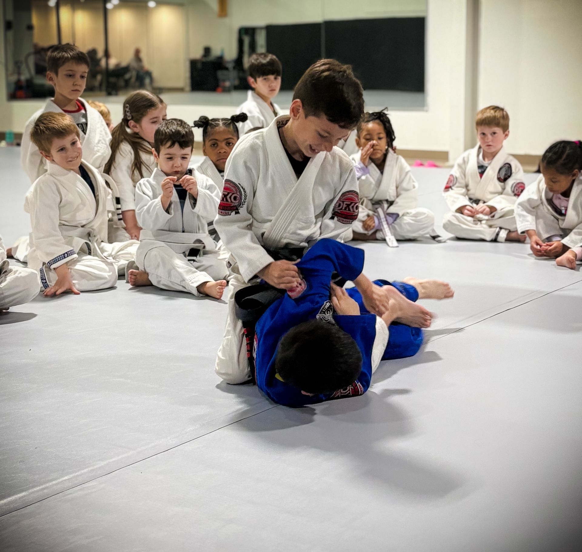 Children in white martial arts uniforms practicing grappling techniques on a mat while classmates observe in a studio.