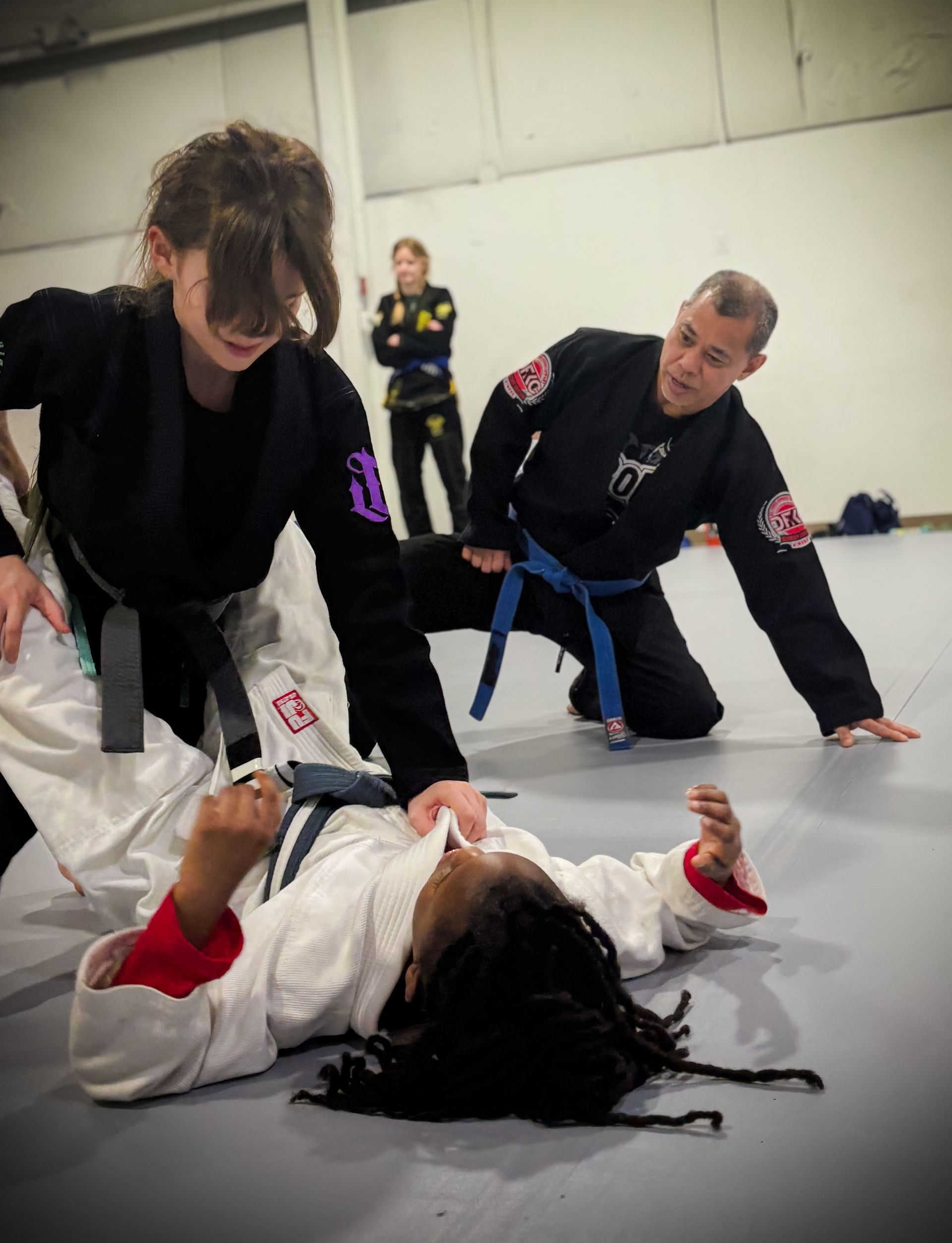 A martial arts instructor watches as two students practice a grappling technique on a gym mat.
