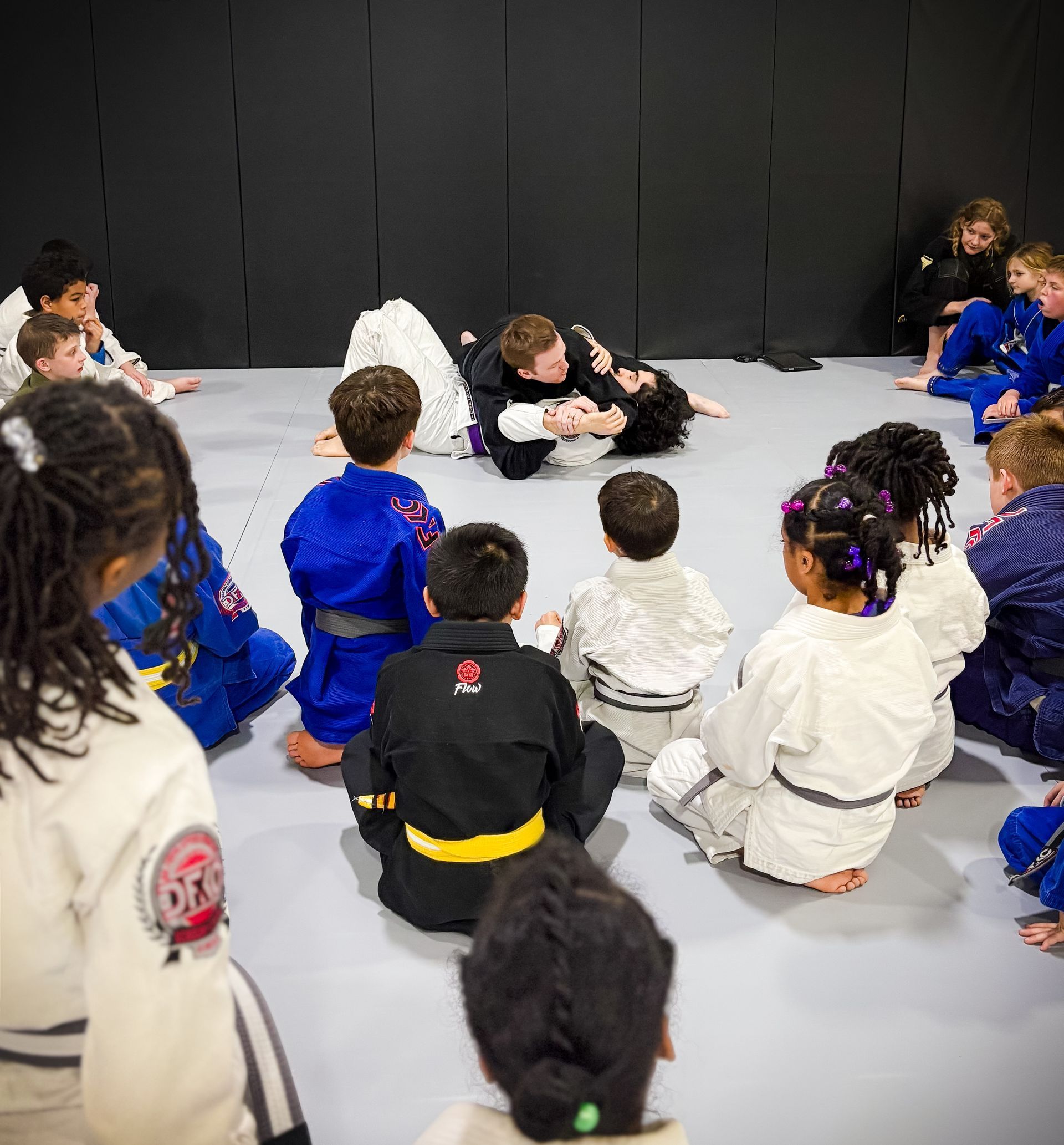 An instructor demonstrates a grappling technique on the floor for a group of children in white and blue martial arts gis.