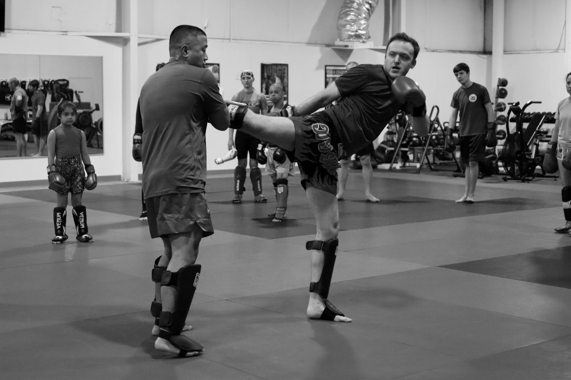 Two sparring partners in a gym training kickboxing, with the fighter on the right delivering a high roundhouse kick.