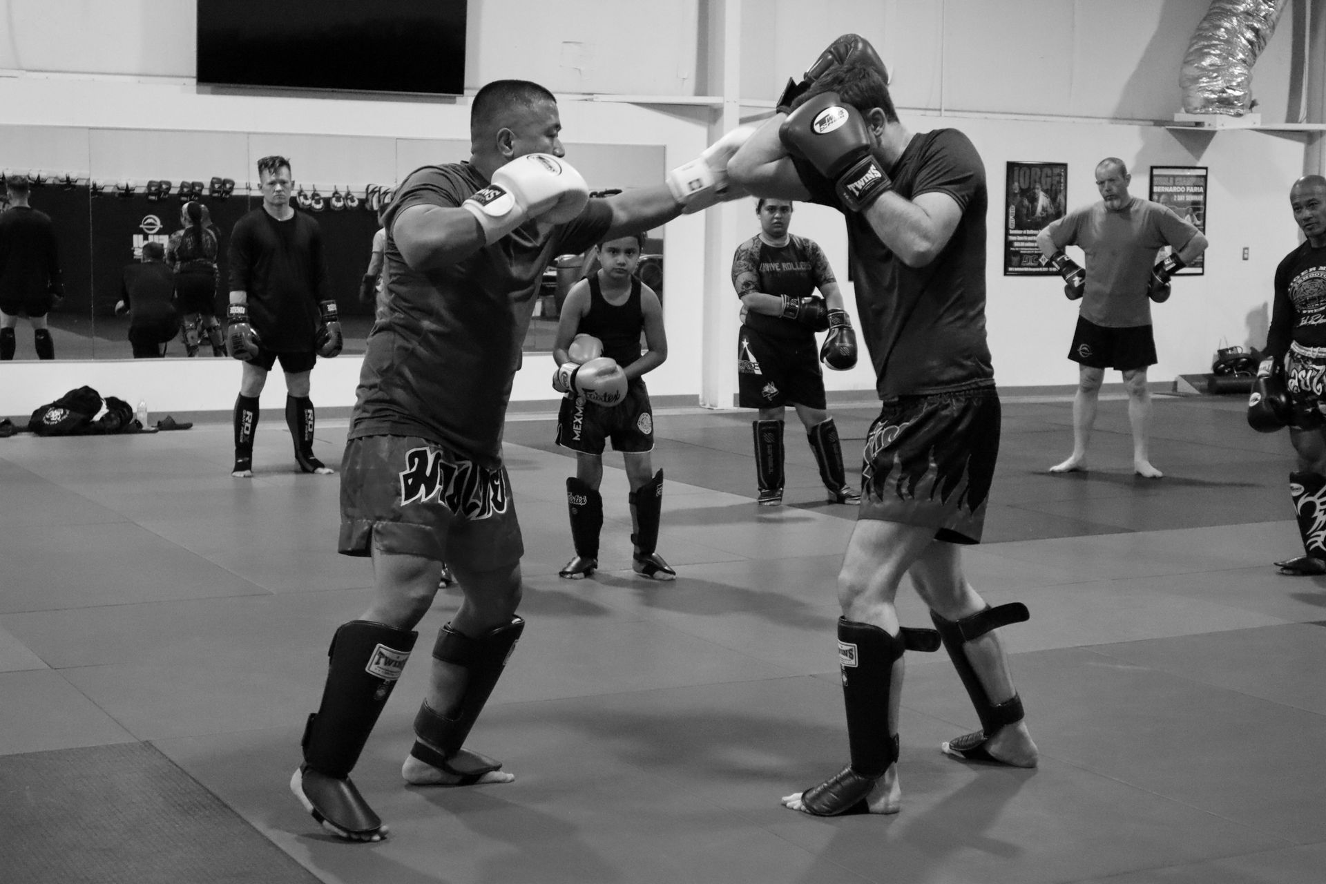 Two sparring partners wearing gloves and shin guards practice Muay Thai techniques in a gym, watched by others nearby.