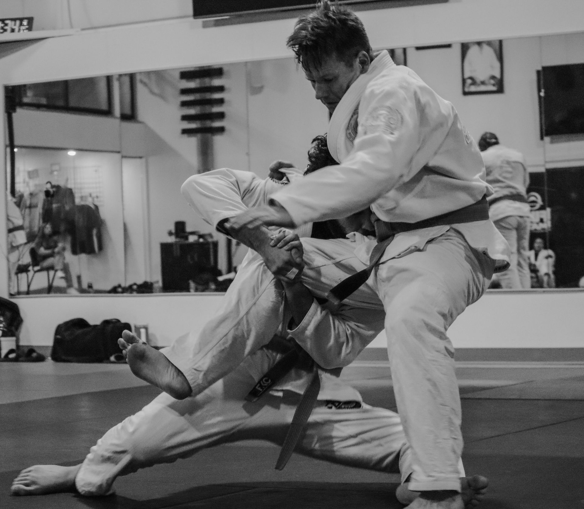 Two people wearing martial arts uniforms grappling on a mat in a training studio, captured in black and white.