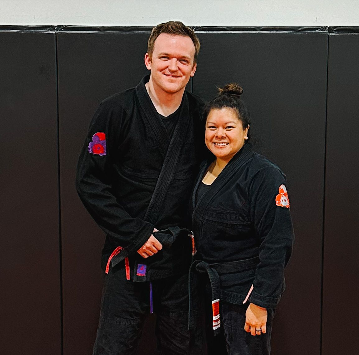 Two smiling martial artists in black gis stand side-by-side against a black wall mat.