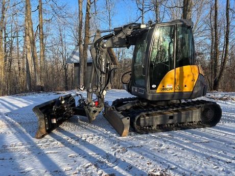 A yellow and black excavator is parked in the snow with trees in the background.