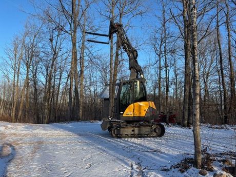 A yellow excavator with fork lift is driving down a snowy road in the woods.