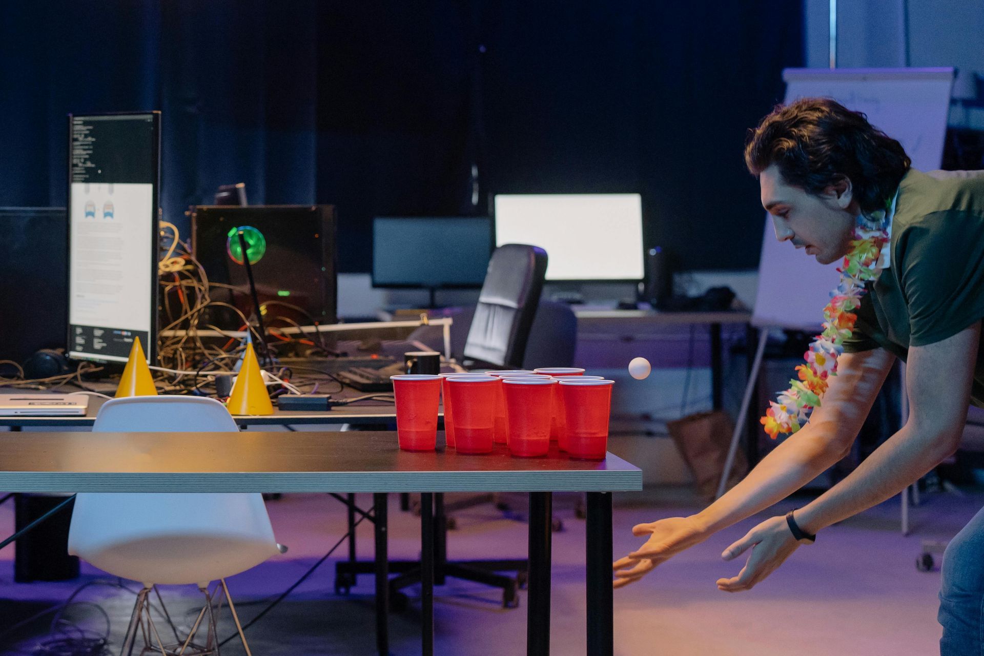 Man in Hawaiian lei, reaching toward red cups on table, playing beer pong in an office.