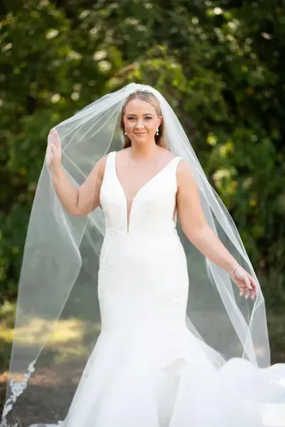 Bride in a white gown and veil standing outdoors, holding her veil with greenery behind her