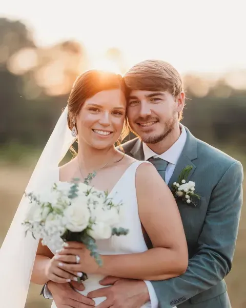 Bride and groom embracing outdoors at sunset, smiling with bouquet and boutonniere.