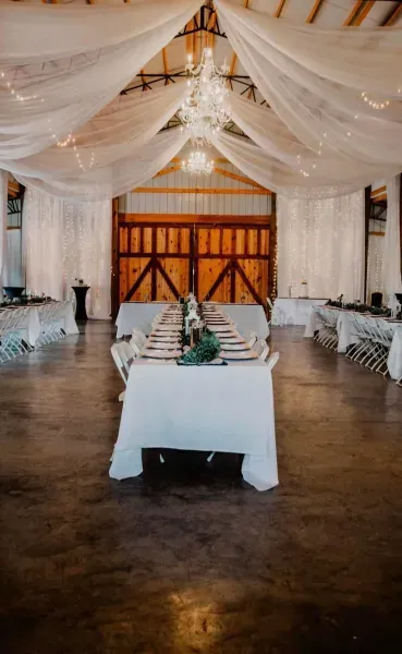 White-draped wedding reception hall with long tables and a central table, viewed from the aisle.