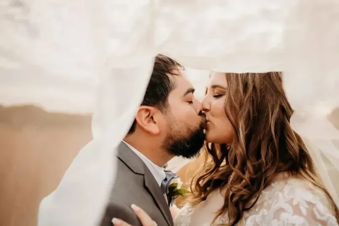 Bride and groom kissing under a white veil outdoors at sunset