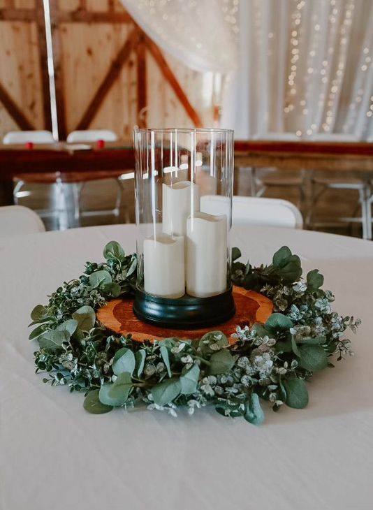 Table centerpiece with white candles in glass holders, surrounded by greenery on a decorated table.