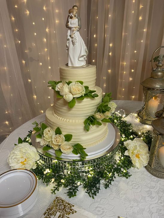 Elegant white wedding cake with floral decorations and bride topper on a glittering table, with twinkling lights backdrop