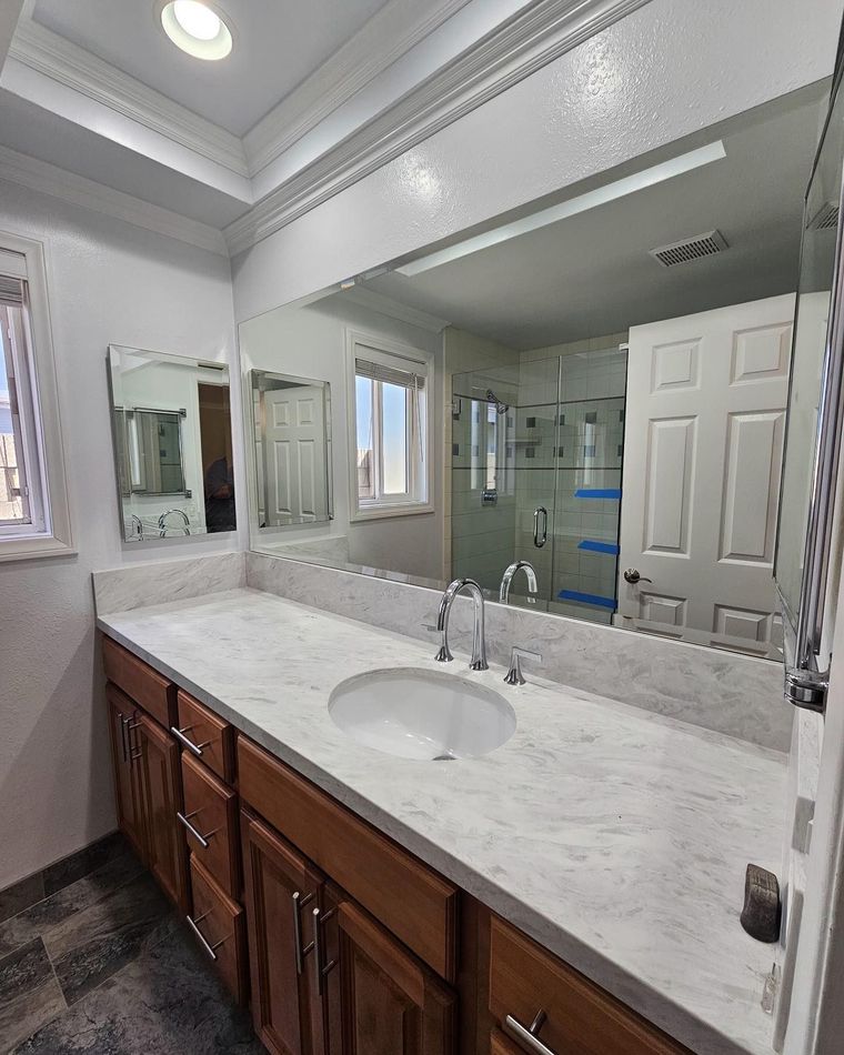 Bright bathroom vanity with large mirror, marble countertop, wooden cabinets, and glass shower in reflection