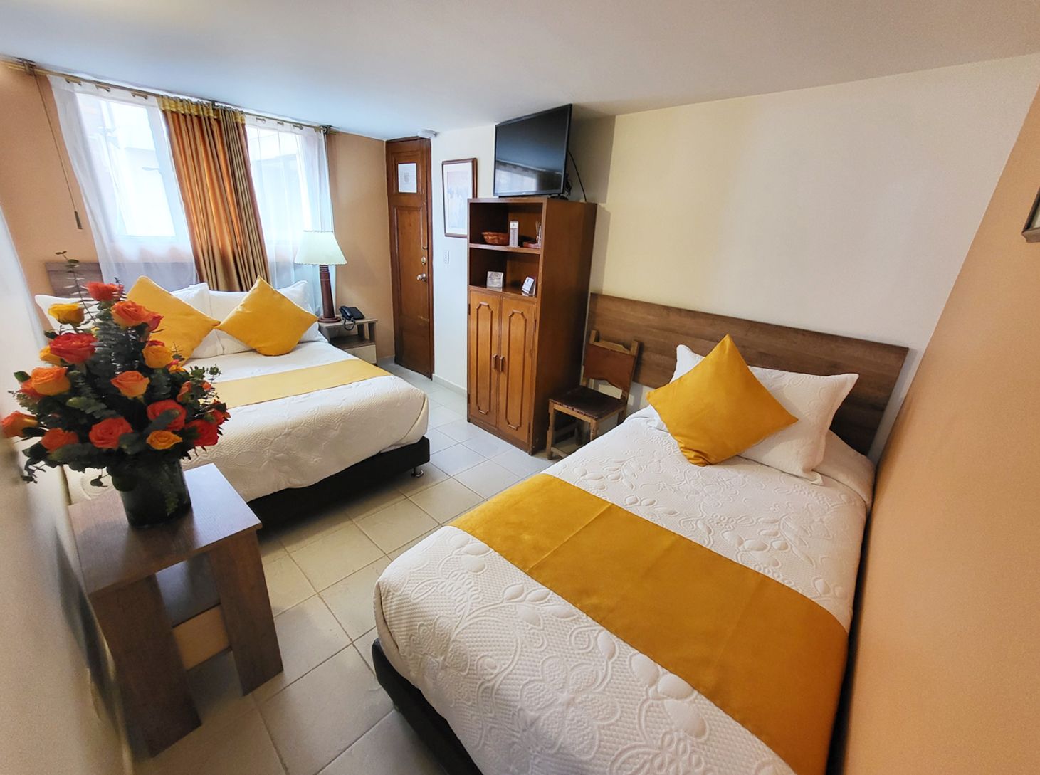 Two beds in a hotel room with yellow accents, a TV, small furniture, and a vase of flowers.