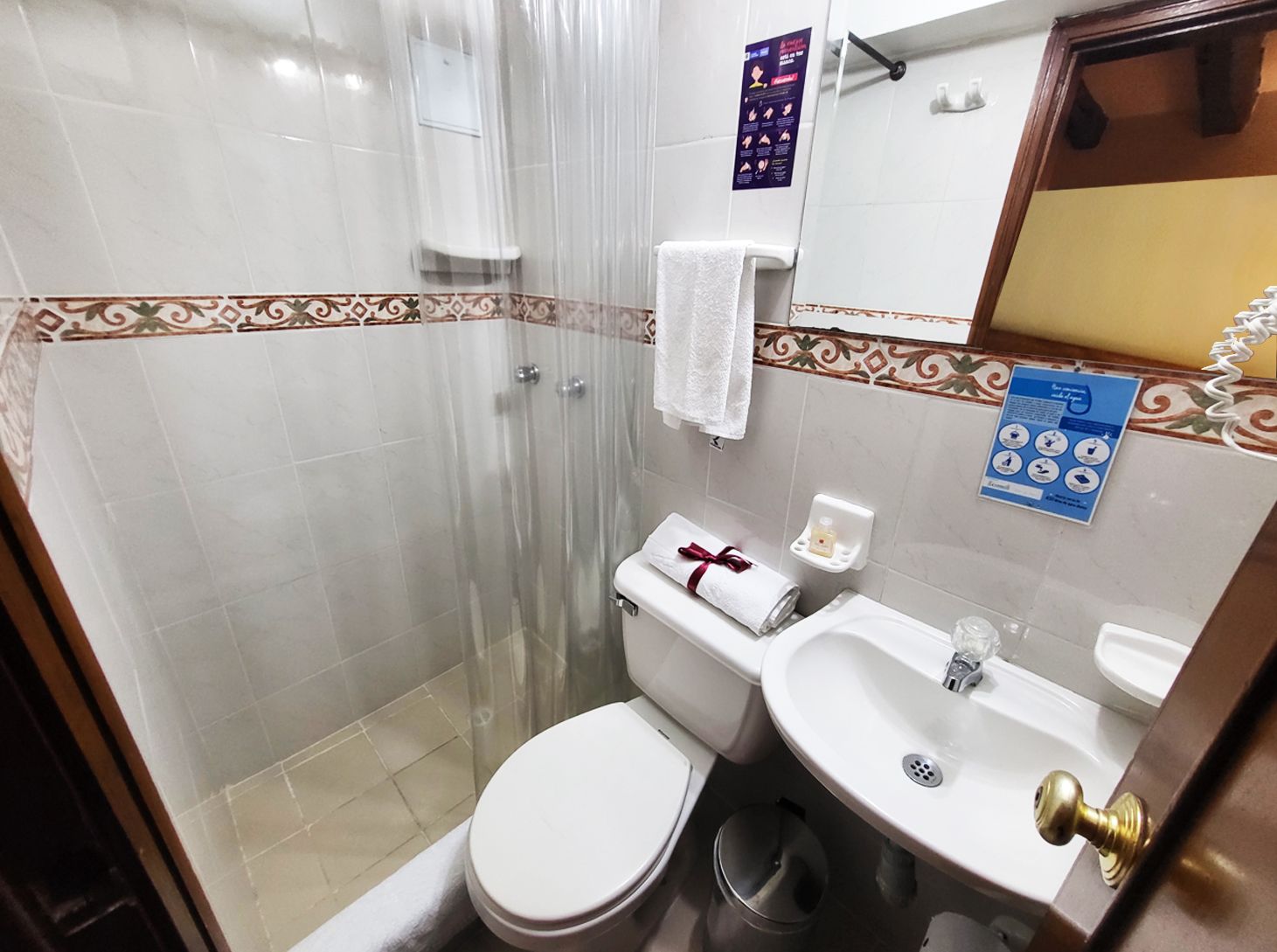 Small hotel bathroom with shower, toilet, sink, and mirror. White and beige tiles.