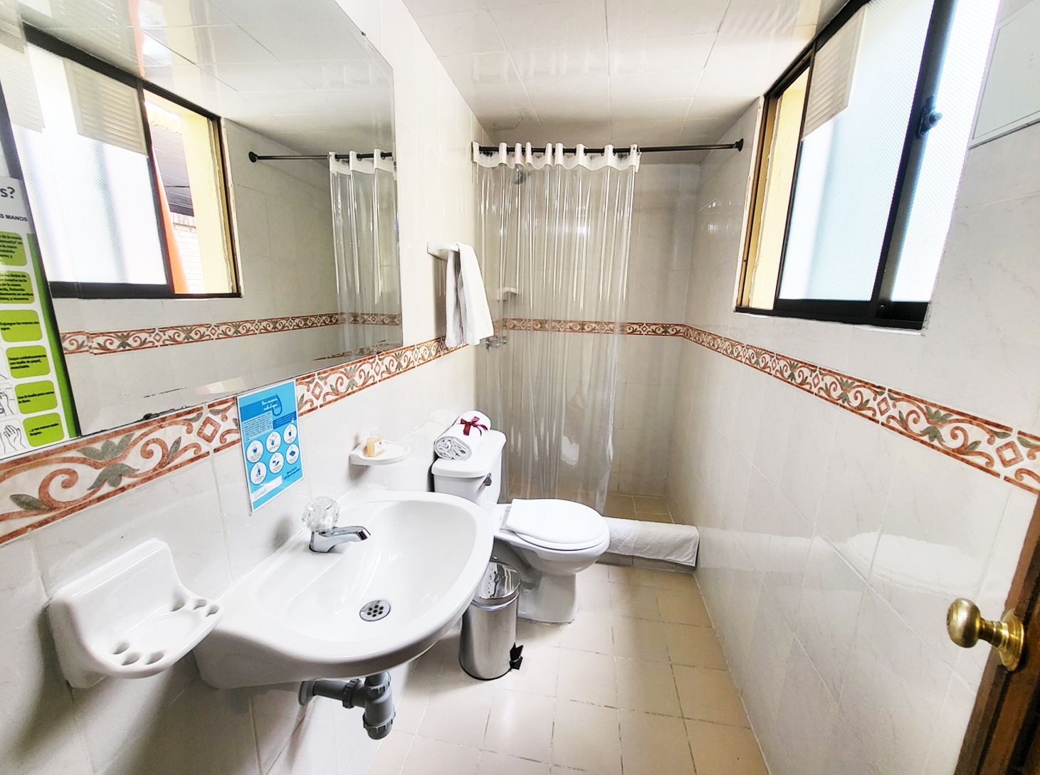 Small, tiled bathroom with sink, toilet, shower curtain, mirror, and two windows.