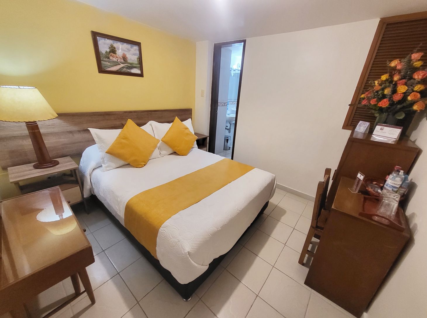 Hotel room with a bed, desk, and bathroom door. Yellow and white linens, floral arrangement on the desk.