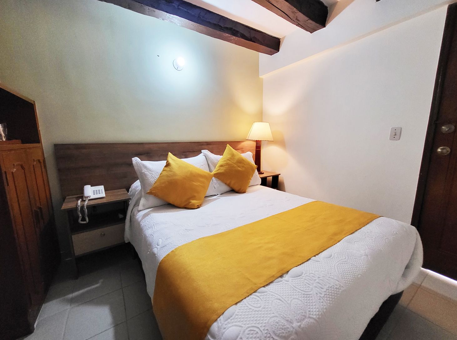 Cozy hotel room with a bed, yellow accents, a nightstand, and dark wooden beams on the ceiling.