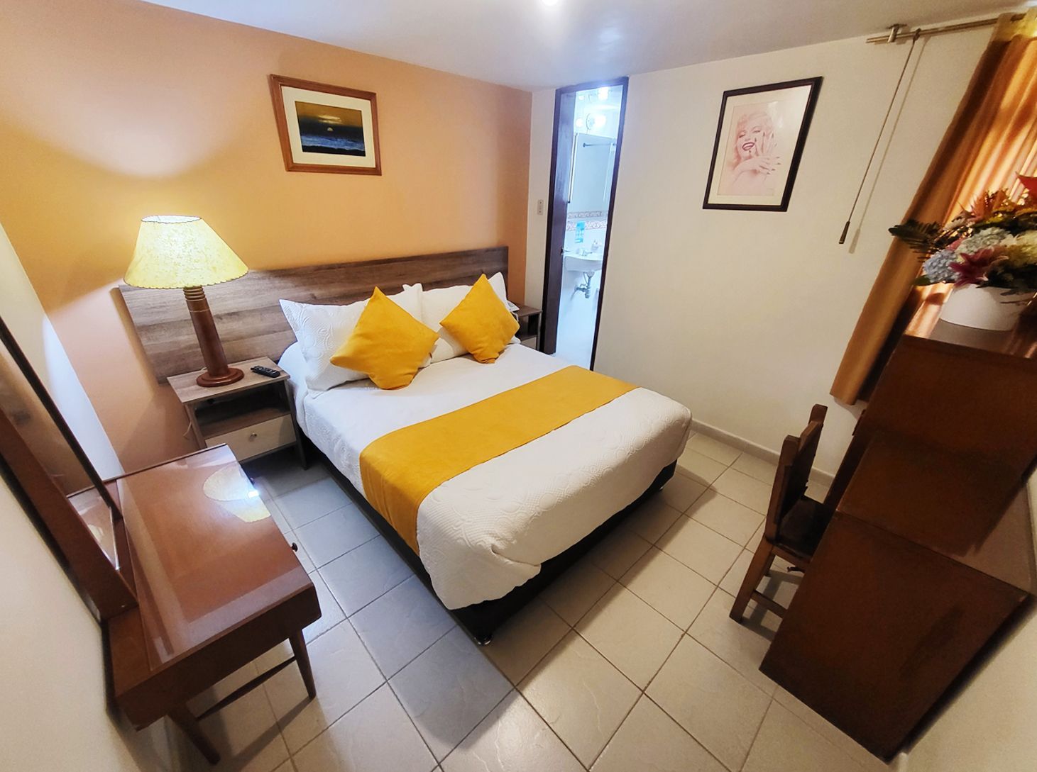 Hotel room with bed, dresser, desk, and lamp. Orange wall with art, floral decorations, tiled floor.